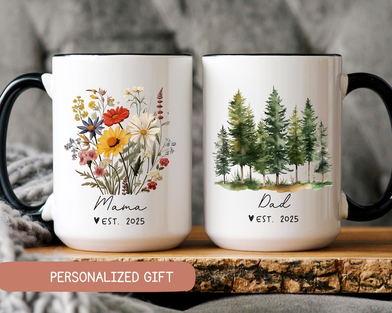 Pode incluir: Duas canecas de cer&acirc;mica branca com al&ccedil;as pretas. A caneca &agrave; esquerda tem um desenho floral e o texto "Mama Est. 2025". A caneca &agrave; direita tem uma cena de floresta em aquarela e o texto "Dad Est. 2025".