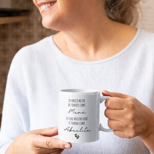 May include: White ceramic mug with black text that reads "Lo unico mejor de tenerte como Mama es que nuestro bebe te tendra como Abuelita" with a pair of baby footprints below the text.