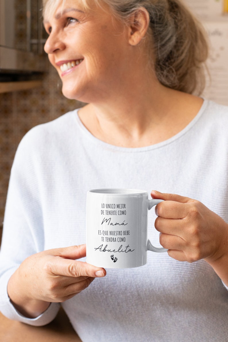 May include: White ceramic mug with black text that reads "Lo unico mejor de tenerte como Mama es que nuestro bebe te tendra como Abuelita" with a pair of baby footprints below the text.