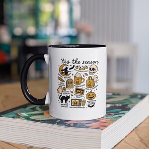 May include: A white ceramic mug with a black handle and rim, decorated with Halloween-themed illustrations and the phrase "'tis the season." The design includes a witch, pumpkins, ghosts, and other spooky elements. The mug is on a stack of books.