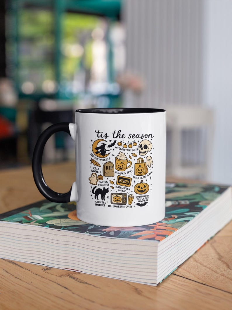 May include: A white ceramic mug with a black handle and rim, decorated with Halloween-themed illustrations and the phrase "'tis the season." The design includes a witch, pumpkins, ghosts, and other spooky elements. The mug is on a stack of books.