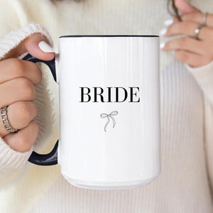 May include: White ceramic mug with a black rim and handle. The mug has the word "BRIDE" printed in black with a small black bow underneath.