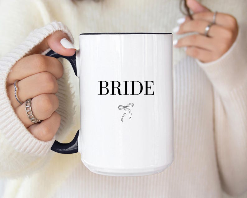 May include: White ceramic mug with a black rim and handle. The mug has the word "BRIDE" printed in black with a small black bow underneath.