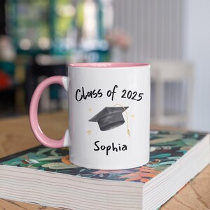 May include: White ceramic mug with a pink rim and handle. The mug features a black graduation cap graphic and the text "Class of 2025" and "Sophia".