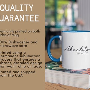 May include: White ceramic mug with a blue rim and handle. The mug has the text "Abuelito EST. 2023" printed on it in black cursive font. The mug is sitting on a stack of books with a floral pattern.