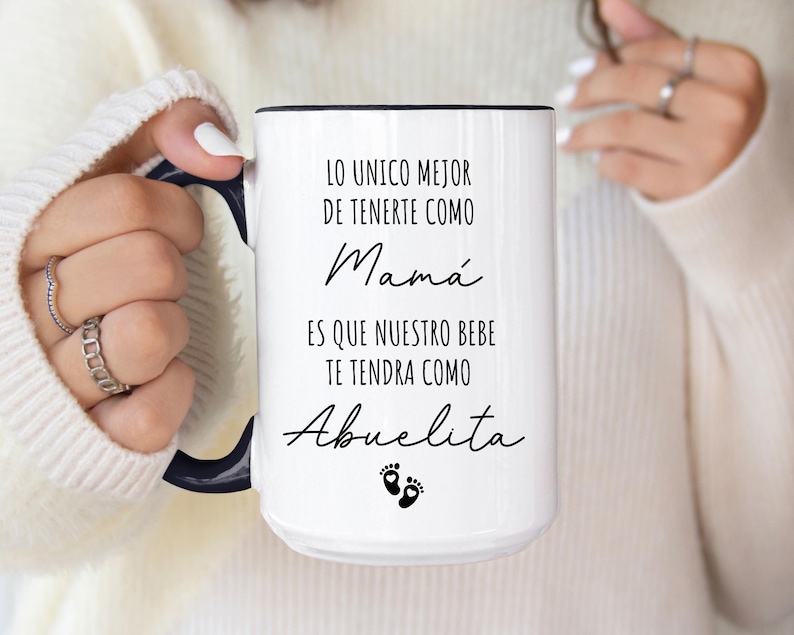 May include: White ceramic mug with a black handle and black text that reads "Lo unico mejor de tenerte como Mama es que nuestro bebe te tendra como Abuelita".