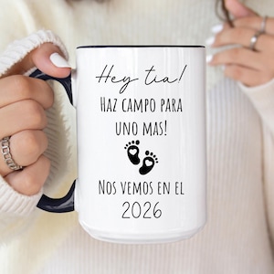 May include: White ceramic mug with a black handle and black text that reads "Hey tia! HAZ CAMPO PARA UNO MAS! NOS VEMOS EN EL 2026". The text is surrounded by two black footprints with hearts.
