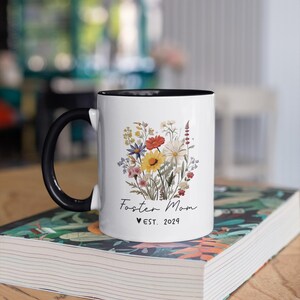 Puede incluir: Taza de cer&aacute;mica blanca con borde y asa negros. La taza presenta un dise&ntilde;o floral con una variedad de flores coloridas y el texto "Foster Mom &hearts; EST. 2024".
