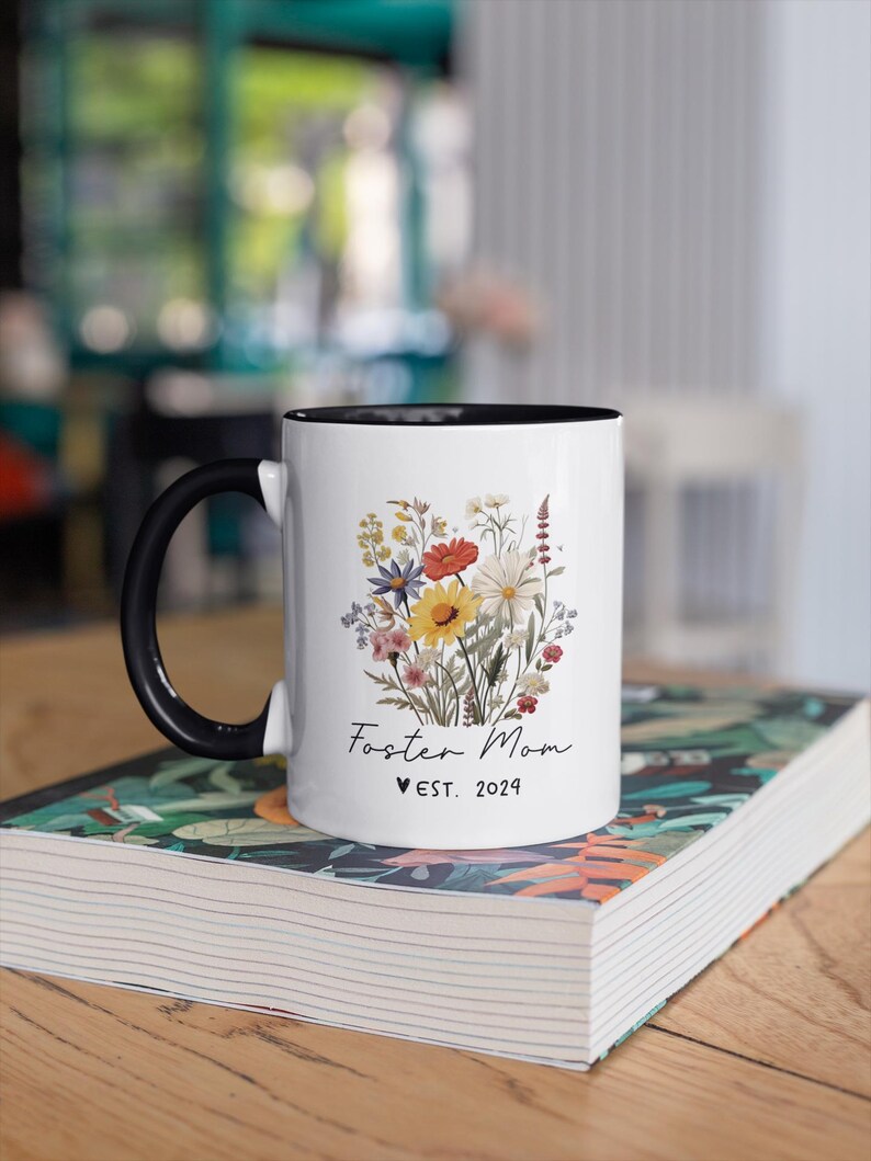 Puede incluir: Taza de cer&aacute;mica blanca con borde y asa negros. La taza presenta un dise&ntilde;o floral con una variedad de flores coloridas y el texto "Foster Mom &hearts; EST. 2024".