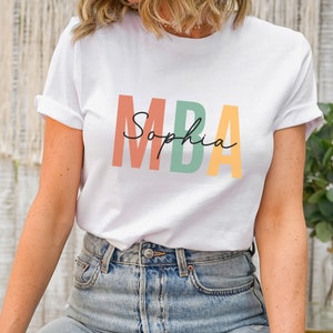 Op de afbeelding: Wit T-shirt met de tekst "MBA" in kleurrijke blokletters en de naam "Sophia" eronder in cursief.