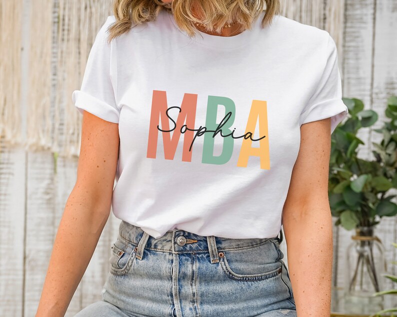 Op de afbeelding: Wit T-shirt met de tekst "MBA" in kleurrijke blokletters en de naam "Sophia" eronder in cursief.