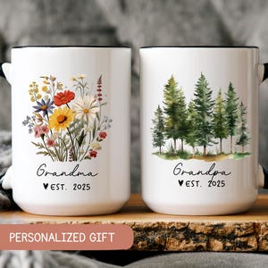 May include: Two white ceramic mugs with black handles and black rims. The mug on the left has a floral design and the text "Grandma Est. 2025". The mug on the right has a forest scene and the text "Grandpa Est. 2025".
