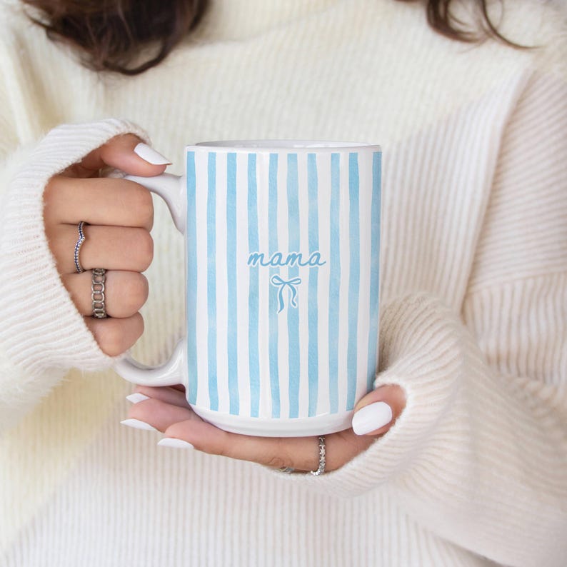 K&ouml;nnte beinhalten: Eine wei&szlig;e Keramiktasse mit vertikalen hellblauen Streifen. Das Wort "mama" ist in Schreibschrift mit einer Schleifenillustration geschrieben. Die Tasse wird von einer Person gehalten, die einen wei&szlig;en Pullover tr&auml;gt.