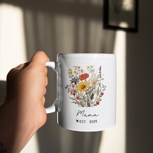 Pode incluir: Caneca de cer&acirc;mica branca com um design floral com uma variedade de flores coloridas. A caneca tem uma al&ccedil;a preta e o texto "Mama &hearts; EST. 2024" impresso na frente.