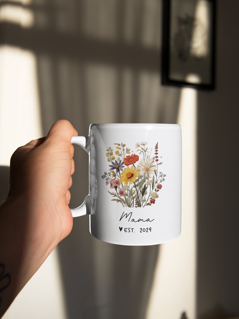 Pode incluir: Caneca de cer&acirc;mica branca com um design floral com uma variedade de flores coloridas. A caneca tem uma al&ccedil;a preta e o texto "Mama &hearts; EST. 2024" impresso na frente.