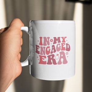 May include: White ceramic mug with a pink and red graphic that says "In My Engaged Era" with a small ring graphic.