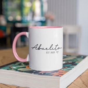May include: White ceramic mug with a pink rim and handle. The mug has the text "Abuelito Est. 2023" in black script.