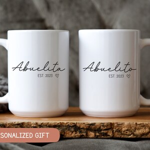 May include: Two white ceramic mugs with black text. The mug on the left says "Abuelita Est. 2023" and the mug on the right says "Abuelito Est. 2023".