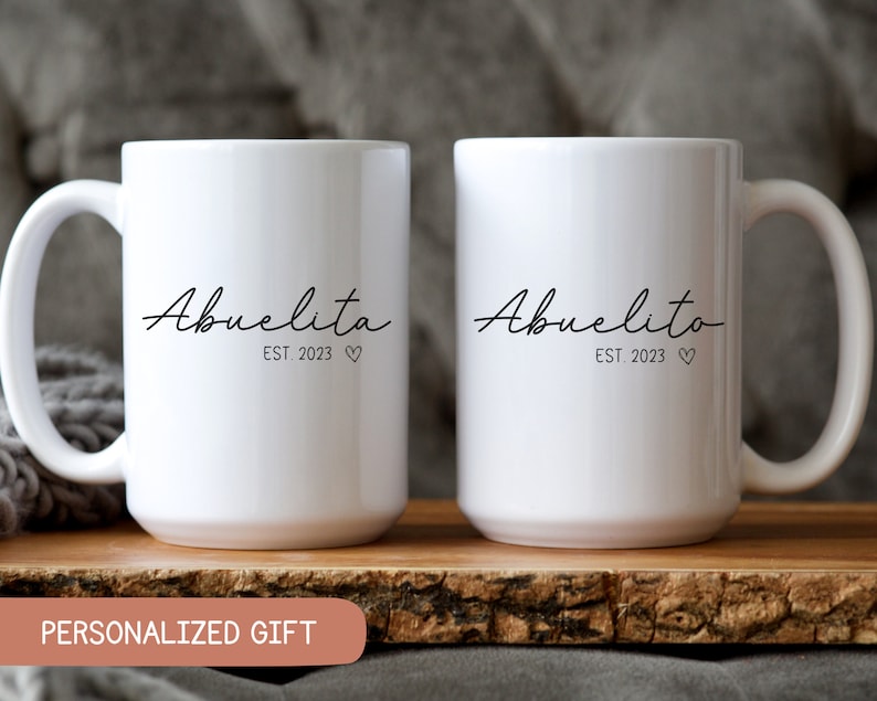 May include: Two white ceramic mugs with black text. The mug on the left says "Abuelita Est. 2023" and the mug on the right says "Abuelito Est. 2023".