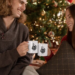 May include: Two white coffee mugs with black skeleton hands giving the middle finger. The mugs say "His" and "Hers" on them. A couple are holding the mugs and smiling at each other in front of a Christmas tree.