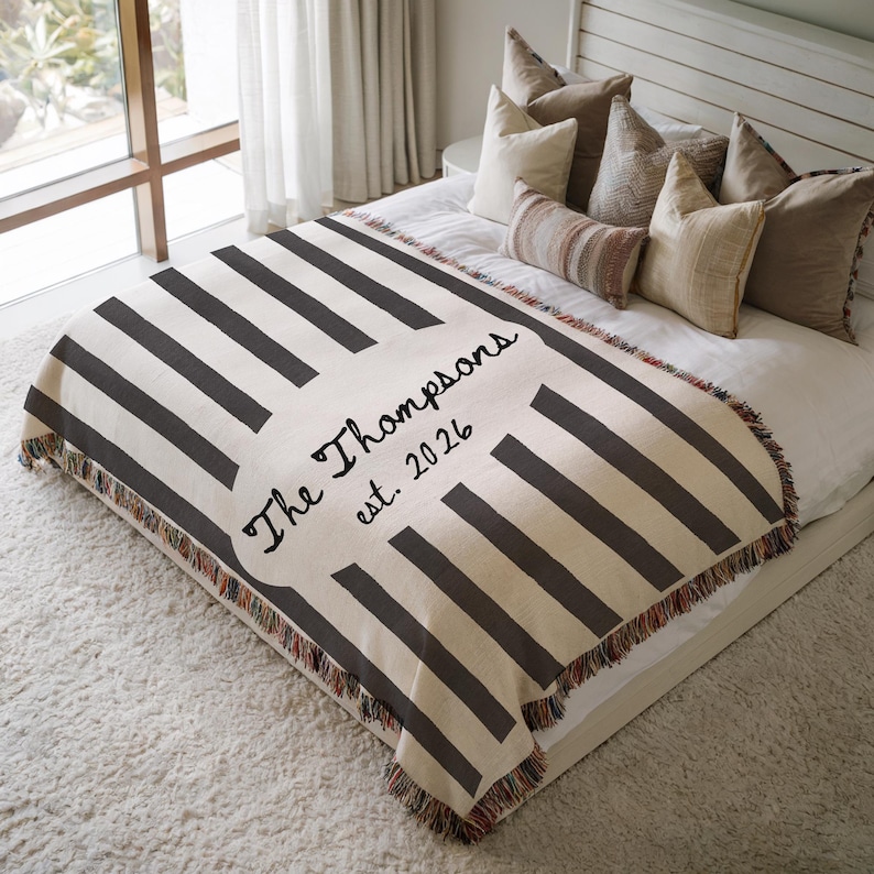 May include: A personalized throw blanket with a cream background and dark gray stripes. The blanket features the text "The Thompsons est. 2026" in a decorative font. The edges are finished with a fringed trim.