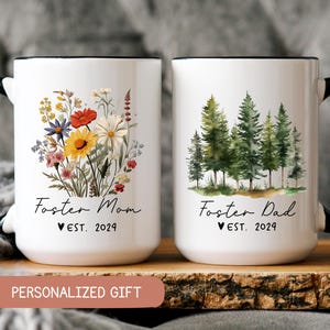 May include: Two white ceramic mugs with black handles and black rims. The mug on the left has a floral design and the text "Foster Mom ♥ EST. 2024". The mug on the right has a watercolor forest design and the text "Foster Dad ♥ EST. 2024".