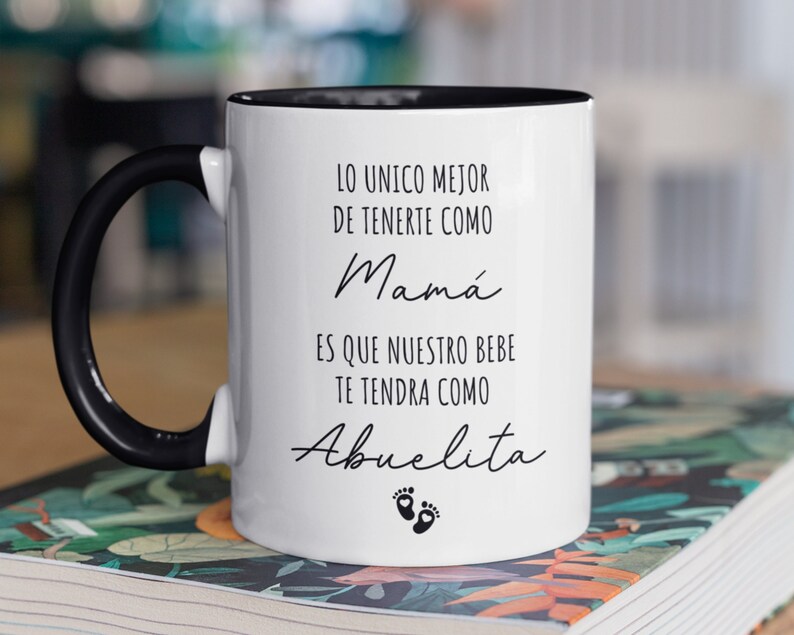 May include: White ceramic mug with a black handle and black rim. The mug has black text that reads "Lo unico mejor de tenerte como Mama es que nuestro bebe te tendra como Abuelita" with two black footprints below the text.