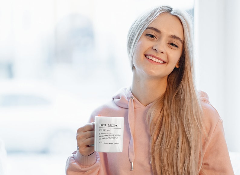 May include: A white ceramic mug with a black inscription that reads "Boss Lady" with a heart symbol. The mug is held by a person wearing a pink hoodie.