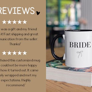May include: White ceramic mug with a black handle and black rim. The mug has the word "BRIDE" printed in black letters with a small black bow underneath. The mug is sitting on a book with a floral pattern.