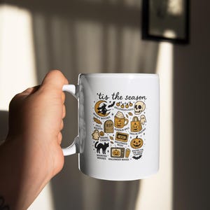 May include: White ceramic mug with a handle, decorated with a Halloween design. The mug displays the text "'tis the season" and Halloween icons such as a witch, pumpkin spice latte, and jack-o'-lanterns. The mug is held by a person.