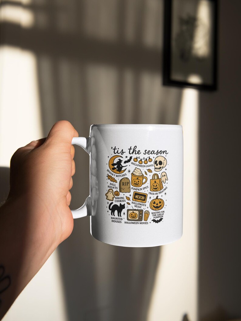 May include: White ceramic mug with a handle, decorated with a Halloween design. The mug displays the text "'tis the season" and Halloween icons such as a witch, pumpkin spice latte, and jack-o'-lanterns. The mug is held by a person.