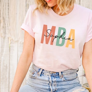 Op de afbeelding: Een roze T-shirt met de tekst "MBA Sophia" in kleurrijke blokletters.