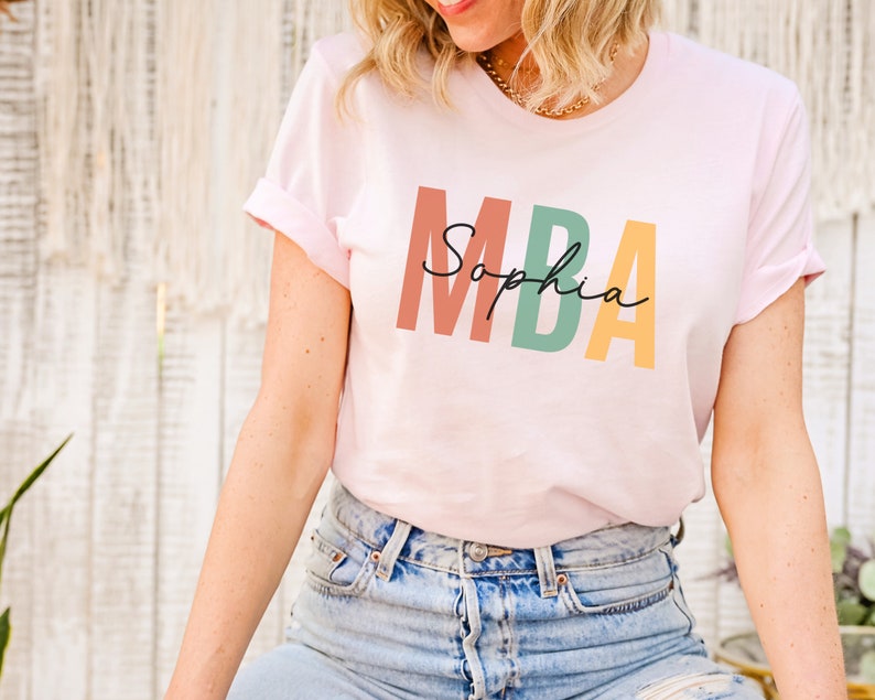 Op de afbeelding: Een roze T-shirt met de tekst "MBA Sophia" in kleurrijke blokletters.