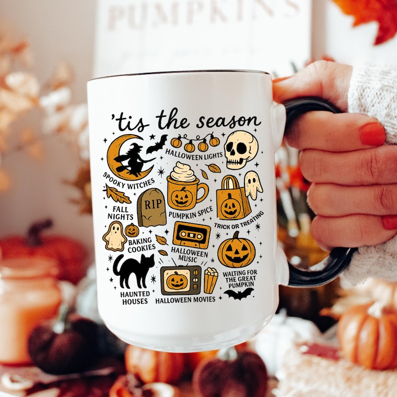 May include: White ceramic mug with a black handle and rim, decorated with Halloween-themed illustrations and text. The design includes a witch on a crescent moon, a skull, pumpkins, and the phrase "'tis the season."
