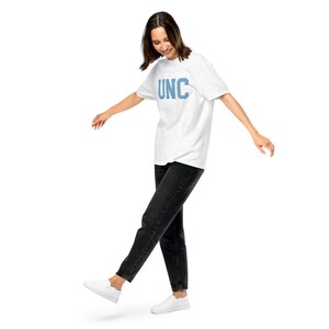 Op de afbeelding: Een vrouw draagt een wit T-shirt met de tekst "UNC" in blauwe letters en een zwarte jeans. Ze loopt met haar linker voet in de lucht en haar rechter voet op de grond.