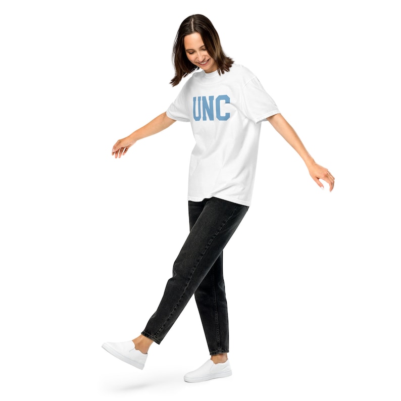 Op de afbeelding: Een vrouw draagt een wit T-shirt met de tekst "UNC" in blauwe letters en een zwarte jeans. Ze loopt met haar linker voet in de lucht en haar rechter voet op de grond.