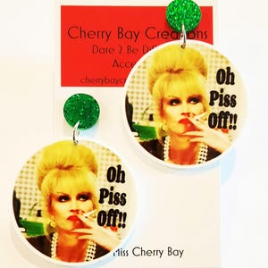 May include: A pair of circular earrings with a woman smoking and the words "Oh Piss Off!!". The earrings have green glitter studs. The earrings are from Cherry Bay Creations, a brand that sells accessories.