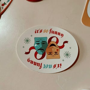 May include: Oval sticker featuring two theatrical masks, one teal and sad, the other tan and smiling, connected by a red ribbon. The text "it's so funny" and "it's not funny" are in red.
