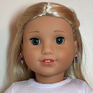 May include: A doll with blonde hair and green eyes wearing small earrings with red and gray beads. The doll is wearing a white shirt and has a neutral expression.