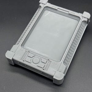 May include: A gray, rectangular device with a recessed screen area and raised edges. The device has a series of buttons and a central circular button. The corners are reinforced with square protrusions. The device is made of a matte material.