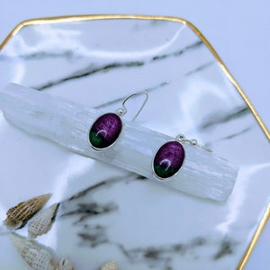 May include: A pair of silver earrings with oval-shaped green and purple gemstones. The earrings are hanging on a white crystal.