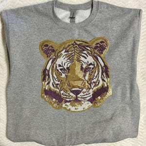 Puede incluir: Una sudadera gris de cuello redondo con un diseño detallado de cara de tigre. La cara del tigre es principalmente dorada con acentos morados y blancos. La sudadera está hecha de un material suave.