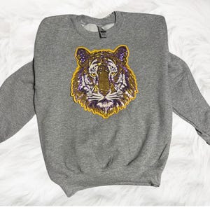 May include: A gray sweatshirt featuring a sequined tiger face design in gold, purple, and white. The tiger's face is centered on the chest of the long-sleeved crew neck sweatshirt. The sweatshirt is made of a soft material.