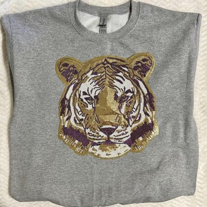 May include: A gray sweatshirt featuring a detailed tiger face design. The tiger's face is embellished with gold, purple, and white sequins. The sweatshirt has a crew neck and long sleeves.