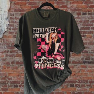 Può includere: Una maglietta verde scuro con una stampa grafica di una donna su uno sfondo a scacchi rosa e nero. Il testo sulla maglietta recita "Hell Yeah I'm the Motherf*ckin Princess".