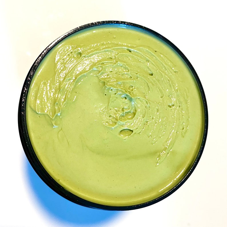 May include: A close-up of a green face mask in a black container. The mask has a smooth, creamy texture.