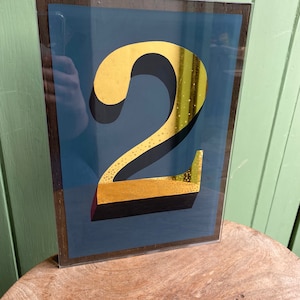 Może przedstawiać: Wydruk w ramie z numerem dwa w kolorze złotym z czarnym obramowaniem na niebieskim tle. Rama jest z ciemnego brązowego drewna.