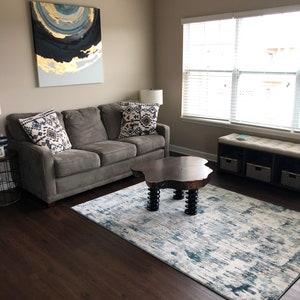 Puede incluir: Salón con un sofá gris, cojines estampados y una mesa de centro de madera. Una alfombra con estampado azul y blanco está en el suelo. Una pintura abstracta cuelga sobre el sofá. La habitación tiene una paleta de colores neutros.