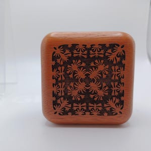 May include: A square wooden box with rounded corners featuring a dark brown laser-engraved floral pattern on the lid. The natural light brown wood grain is visible across the surface, contrasting with the intricate, symmetrical botanical design.