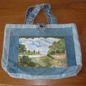 May include: A denim tote bag with a landscape scene featuring cows grazing in a field. The bag has denim handles and a button closure. The landscape scene is framed by a light blue denim border.